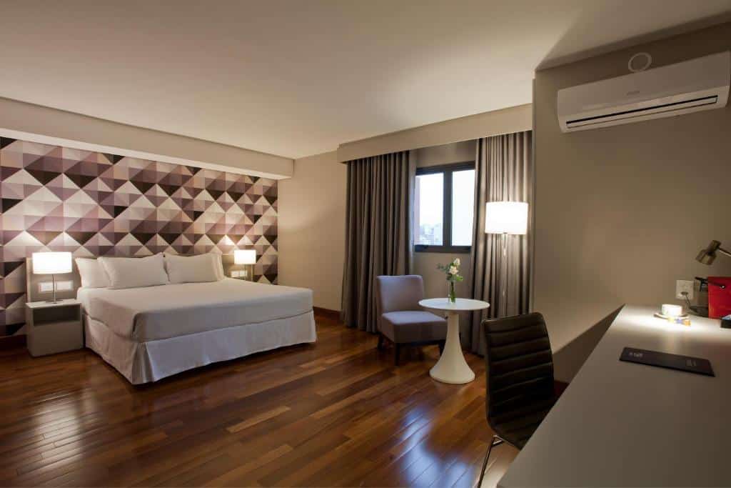 Quarto do NH Curitiba The Five. Uma mesa de trabalho com um ar-condicionado em cima do lado direito. No meio do quarto, no fundo, uma mesa com uma cadeira e a janela do quarto. No canto esquerdo a cama de casal com uma luminária e uma cômoda de cada lado. Foto para ilustrar post sobre hotéis em Curitiba.
