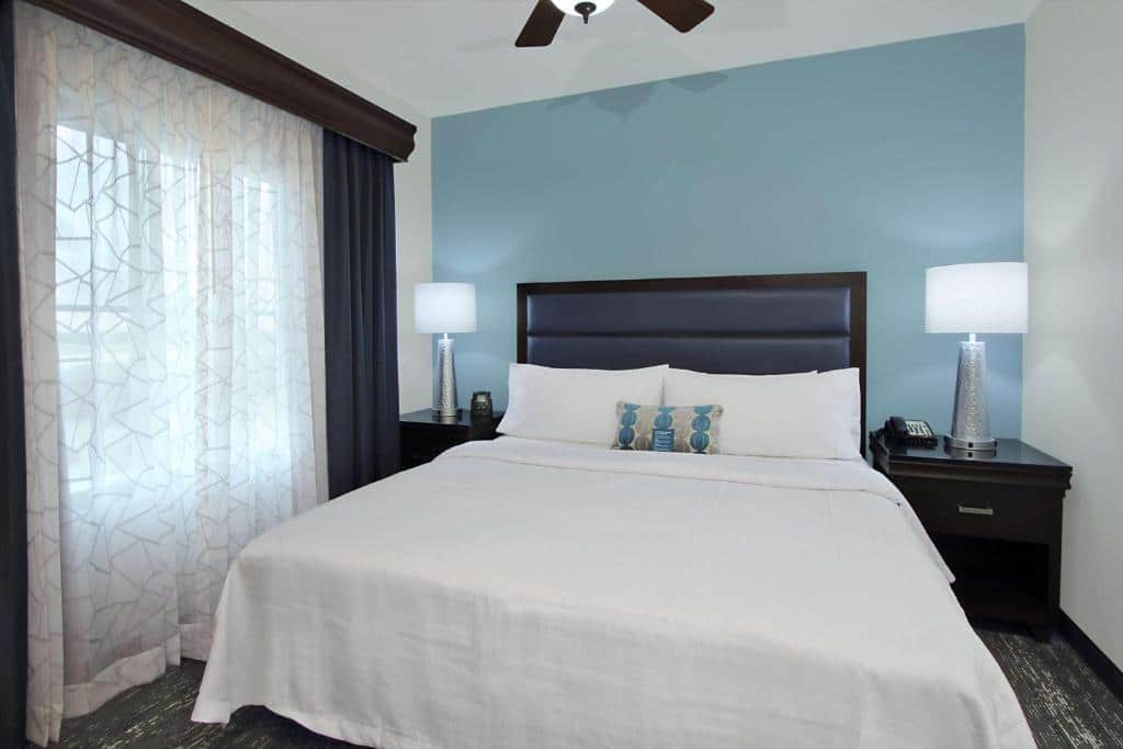 Uma cama bem arrumada do Homewood Suites by Hilton Miami - Airport West
com lençóis brancos, ladeada por duas mesas de cabeceira com luminárias, em um quarto com uma parede azul e uma janela com cortinas transparentes. Representa hotéis em Miami.