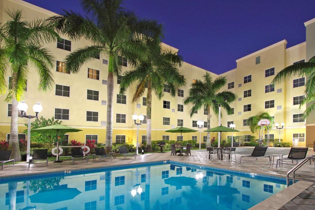 Vista noturna de um pátio do Homewood Suites by Hilton Miami - Airport West
com uma piscina iluminada, palmeiras, espreguiçadeiras e uma cesta de basquete.