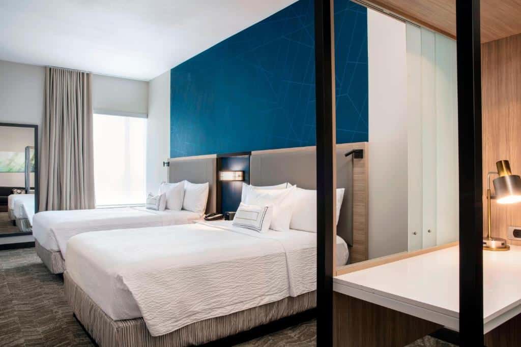 Quarto do SpringHill Suites by Marriott Miami Doral com duas camas de casal, parede com detalhes em azul, escrivaninha com luminária e uma grande janela com cortinas bege. Representa hotéis em Miami.