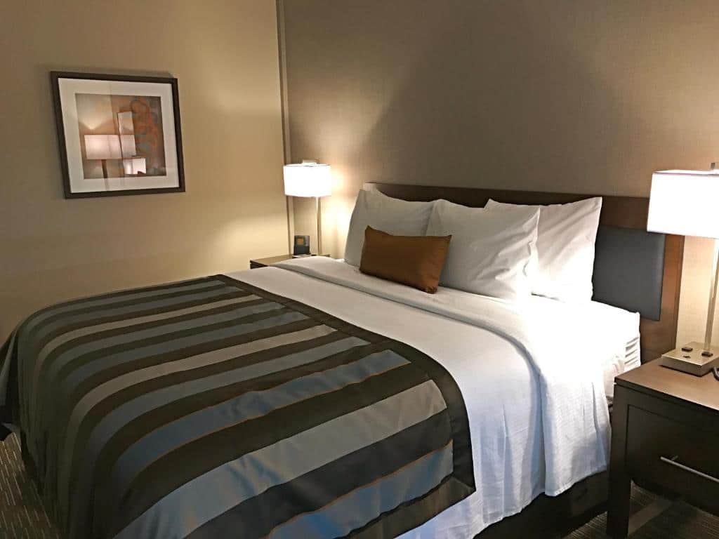 Uma cama bem arrumada do Wingate by Wyndham Miami Airport com roupa de cama listrada, duas mesas de cabeceira com luminárias e uma pintura abstrata emoldurada na parede. Representa hotéis em Miami.