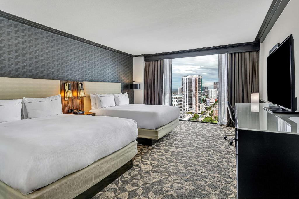 Quarto do Hilton Miami Downtown com duas camas grandes, uma TV montada na parede, uma escrivaninha e uma grande janela mostrando o horizonte da cidade com edifícios altos.