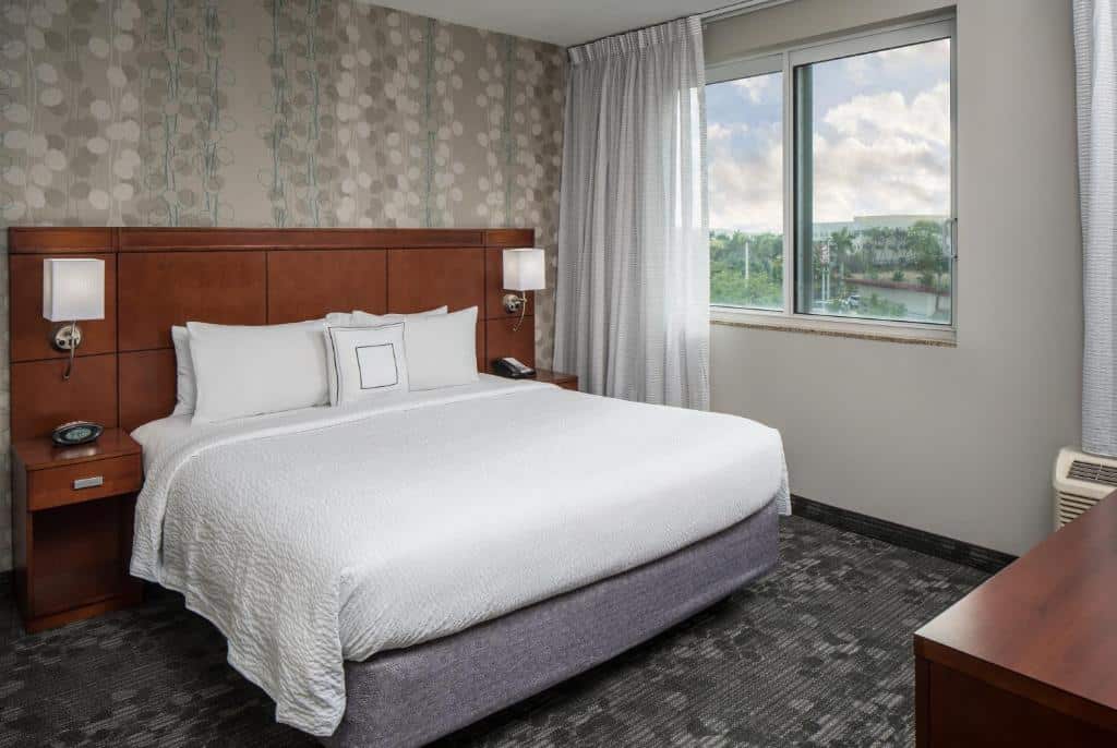 Um quarto do Courtyard by Marriott Miami West/FL Turnpike com uma cama king-size, cabeceira de madeira, dois abajures, uma janela com vista e papel de parede estampado. Representa hotéis em Miami.