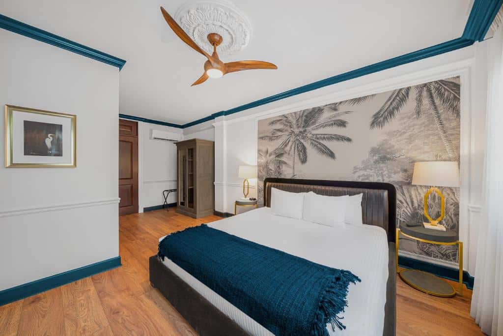 Um quarto do Hotel St. Michel
com uma cama grande, detalhes em azul, papel de parede de palmeira, ventilador de teto e piso de madeira. Um guarda-roupa e luminária estão presentes. Representa hotéis em Miami.