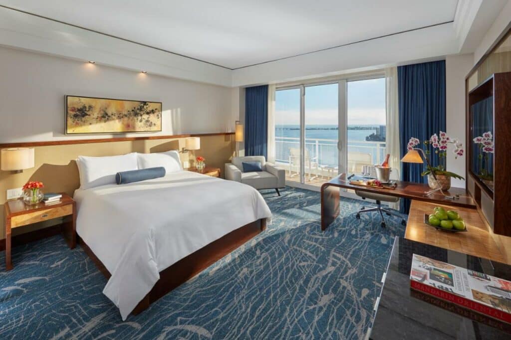 Quarto do Mandarin Oriental, Miami com cama king-size, carpete azul, janela grande com vista para a água, escrivaninha, poltrona e plantas decorativas. Representa hotéis em Miami.