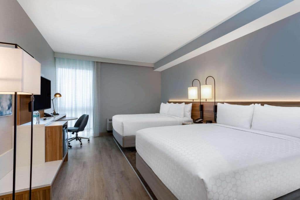 Quarto do Wyndham Garden com duas camas de casal, roupa de cama branca e design moderno. Uma escrivaninha com uma cadeira e uma TV estão posicionadas contra uma parede. 