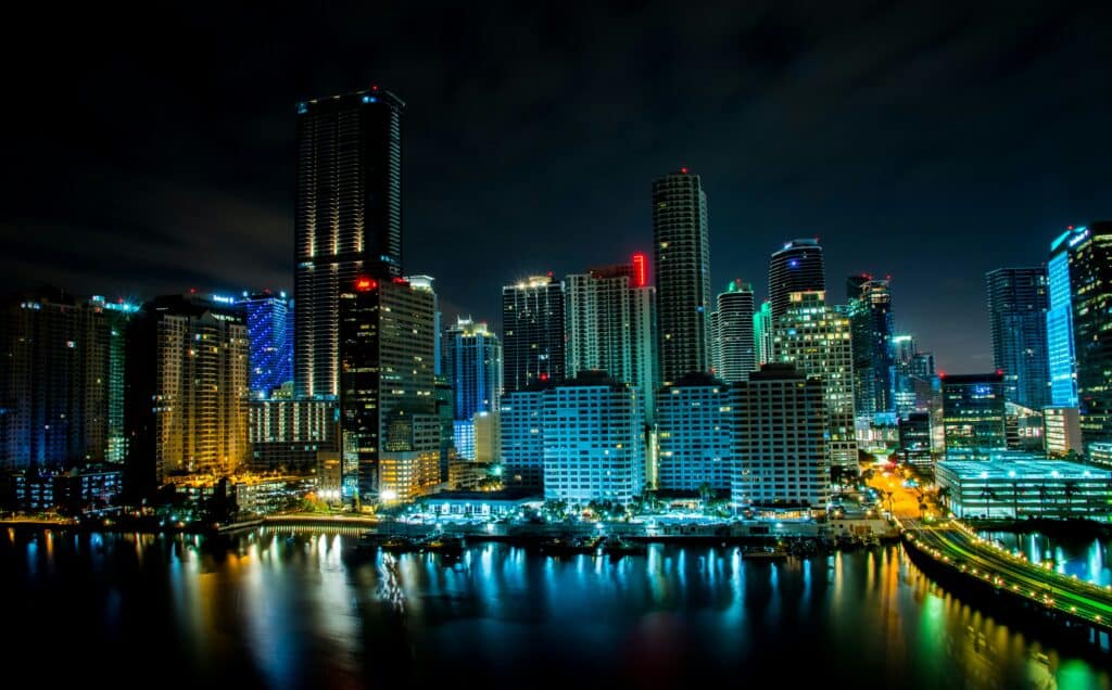 Vista noturna do horizonte de Miami com arranha-céus iluminados refletindo em um rio, apresentando vários estilos arquitetônicos. Representa seguro viagem Miami.