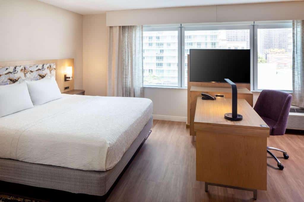 Quarto do Hampton Inn & Suites by Hilton Miami Downtown/Brickell com cama grande, escrivaninha de madeira com TV, cadeira de escritório e janela com vista para a cidade. O quarto tem decoração em cores claras. 