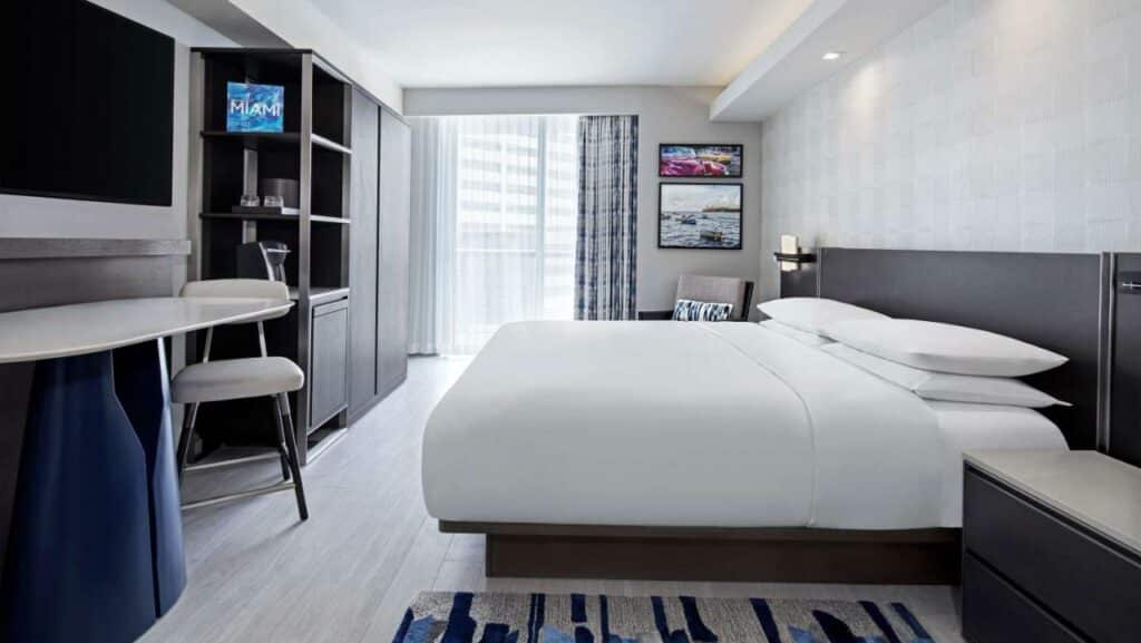 Quarto do Hyatt Centric Brickell com cama grande, escrivaninha, cadeira, TV e varanda. O quarto tem decoração minimalista e esquema de cores claras.