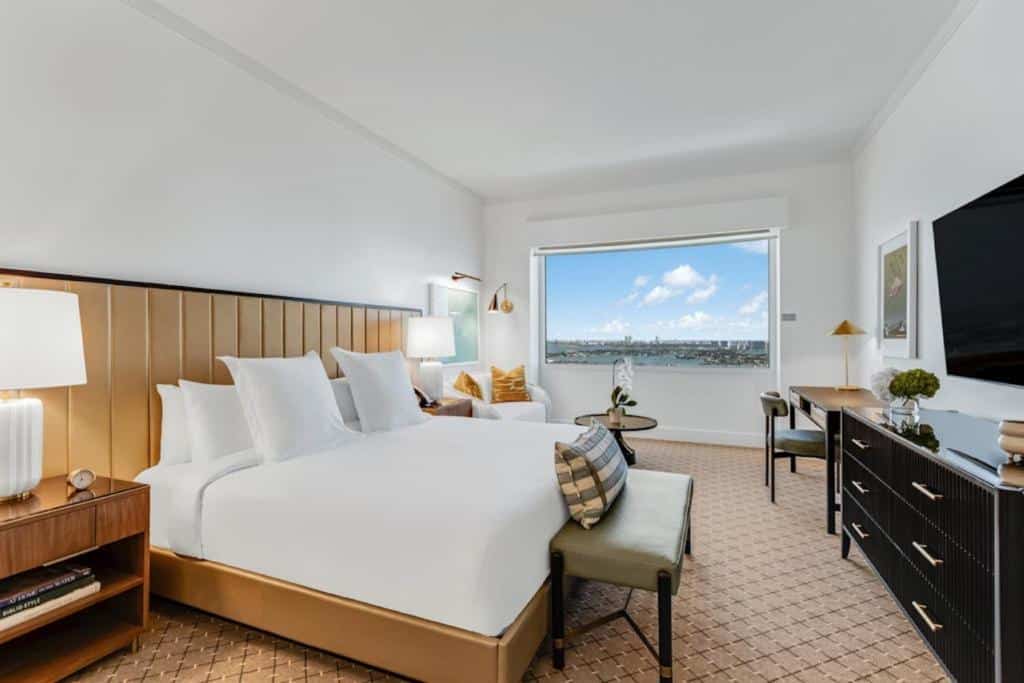 Quarto do InterContinental Miami com cama grande, escrivaninha e vista da janela para um rio e paisagem urbana. Tons neutros e decoração moderna são destaques. Representa hotéis em Miami.