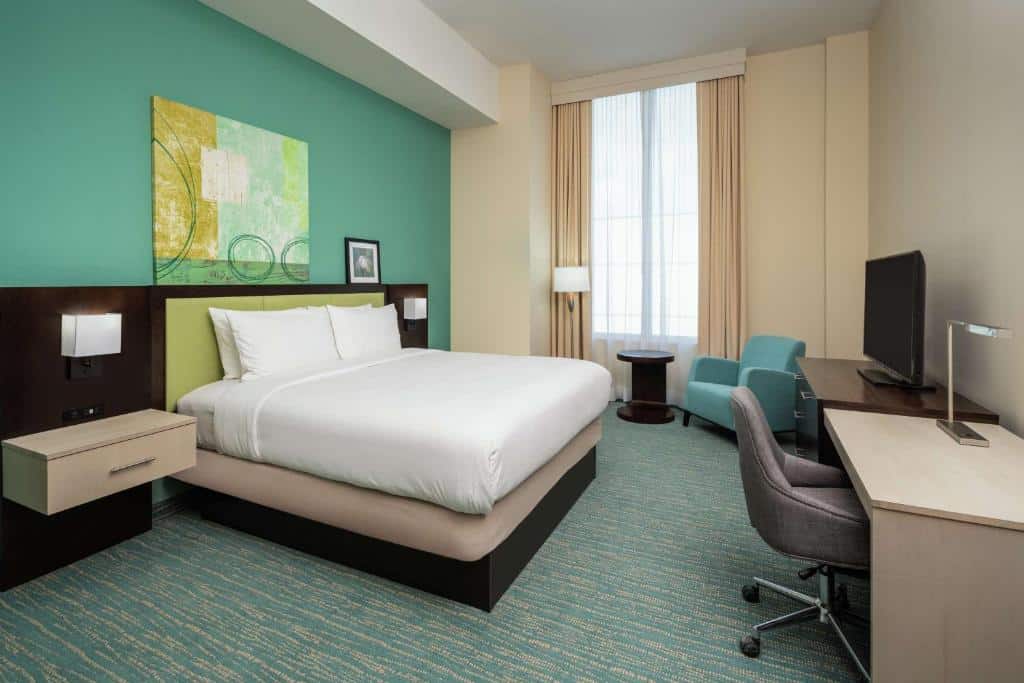 Quarto do DoubleTree by Hilton Miami Doral com uma cama de casal, paredes verdes e bege, uma escrivaninha e cadeira, uma poltrona azul-petróleo, uma luminária de chão e uma janela grande. Representa hotéis em Miami.