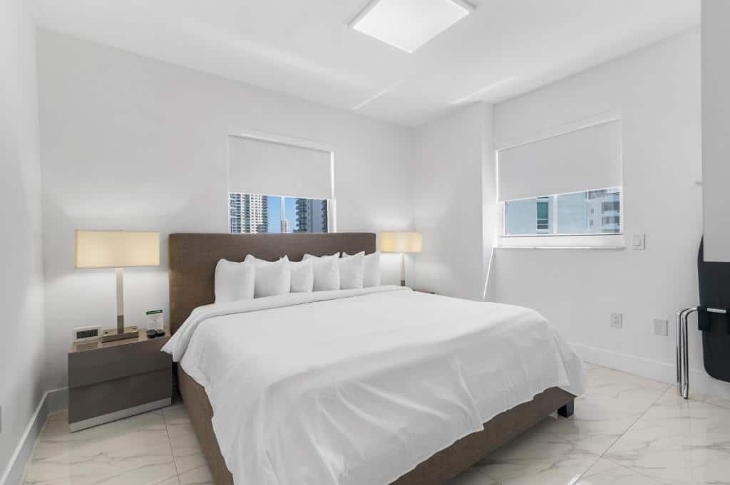 Quarto minimalista do Fortune House Hotel Suites com uma cama grande, lençóis brancos, duas mesas de cabeceira com luminárias e duas janelas com vista para a cidade. 