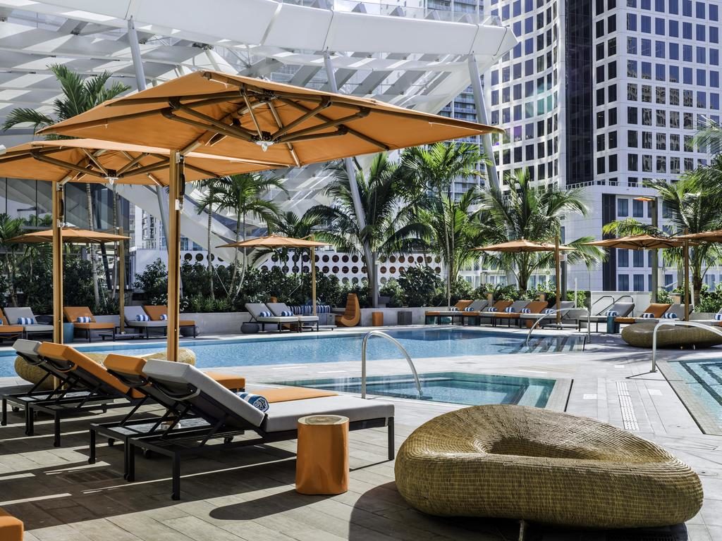 A imagem mostra a área da piscina no East Miami, um dos hotéis em Miami Beach indicados. Há espreguiçadeiras com almofadas laranjas e brancas ao redor da piscina, cobertas por grandes guarda-sóis laranja. Atrás das espreguiçadeiras, há vegetação tropical com palmeiras e arbustos. Em segundo plano, grandes edifícios modernos de vidro e metal compõem o cenário, sugerindo uma localização central em uma cidade grande. A atmosfera é sofisticada e relaxante, ideal para lazer.