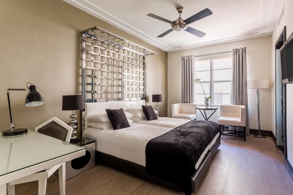 A imagem mostra um quarto de hotel elegante e bem decorado no Waldorf Towers. Há duas camas de solteiro com roupa de cama branca e colchas pretas, decoradas com almofadas pretas. Acima das camas, uma cabeceira decorativa em metal com design geométrico adiciona um toque sofisticado. À esquerda, há uma mesa de trabalho branca com uma luminária moderna. Perto da janela, que tem cortinas cinza, há duas poltronas brancas com uma mesa de vidro entre elas, adornada com uma orquídea. No teto, um ventilador de teto moderno completa o ambiente aconchegante e funcional.