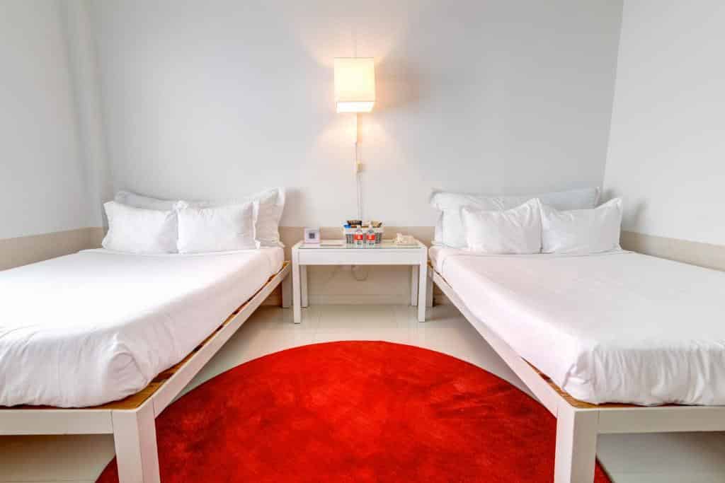 A imagem mostra um quarto simples e moderno com duas camas de solteiro no Townhouse Miami Beach. As camas têm roupa de cama branca, criando um ambiente limpo e minimalista. Entre as camas, há uma mesa de cabeceira branca com alguns itens, como um telefone e livros. Acima da mesa, uma lâmpada de parede ilumina o espaço. No chão, há um tapete redondo vermelho que adiciona um toque de cor vibrante ao ambiente predominantemente branco. As paredes são brancas, reforçando a sensação de simplicidade e clareza no design do quarto.