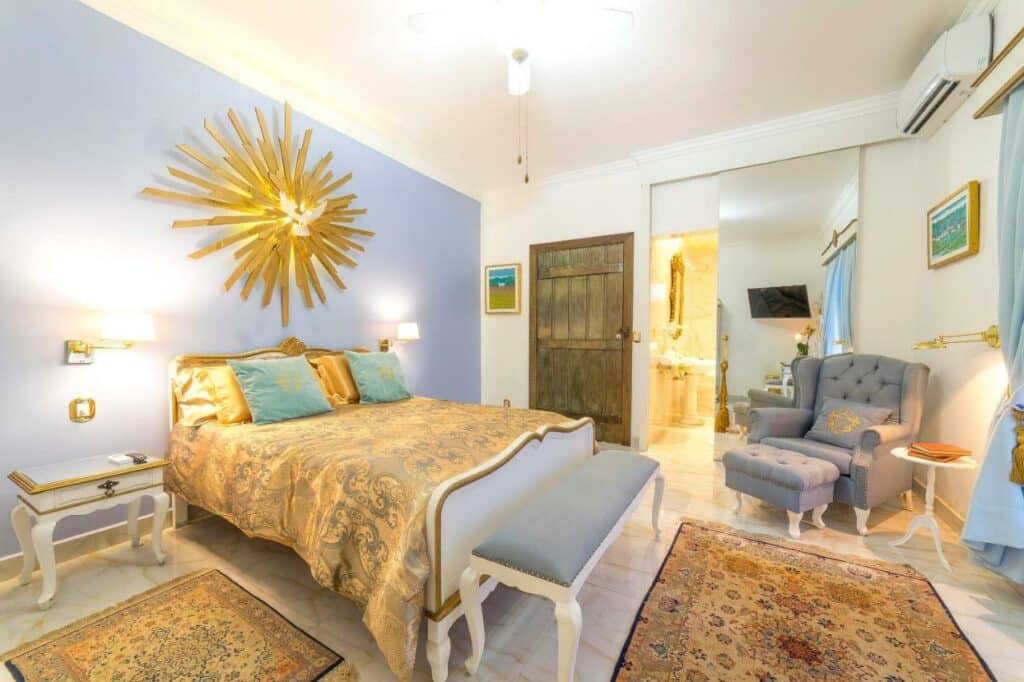 Quarto amplo na Casa Nostra Boutique Hotel com uma decoração bem colonial em tons de dourado, azul e branco, com uma cama de casal, uma poltrona, um ar-condicionado, uma janela ampla, e outros itens de decoração.