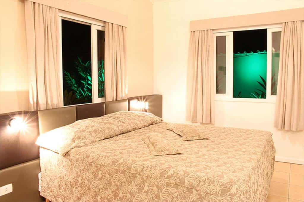 Quarto do Paraty Bungalows Bar E Hotel com uma cama de casal e duas janelas com cortinas, tudo em tons de branco e bege, para representar hotéis em Paraty.
