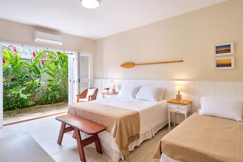 Quarto espaçoso na Casa Clara Paraty com uma cama de casal e uma de solteiro, com uma varanda térrea quando vista para algumas plantas, o quarto é decorado em tons de bege, branco e madeira, para representar hotéis em Paraty.
