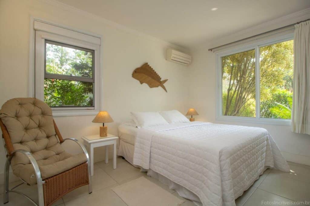 Quarto amplo na Maris Paraty com uma janela com cortina, um ar-condicionado, uma cama de casal com roupas de cama branca, uma mesinha de cabeceira com um abajur, uma poltrona e um quadro de peixe na parede, para representar hotéis em Paraty.