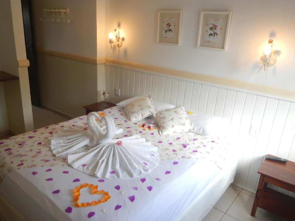 Quarto no Recanto da Praia com uma cama de casal com toalhas e pétalas de rosa, na parede há dois abajures e dois quadrinhos.