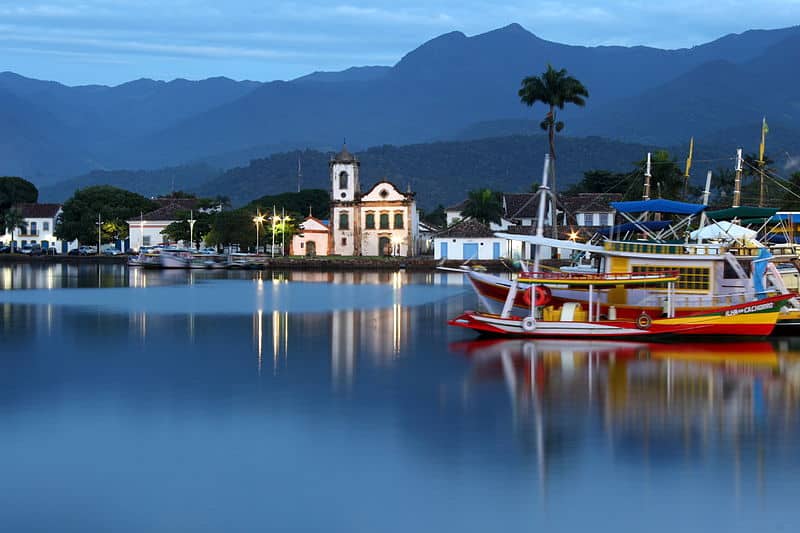 Barcos estacionados no cais de Paraty com montanhas ao fundo e, na beira do cais, algumas construções históricas.
