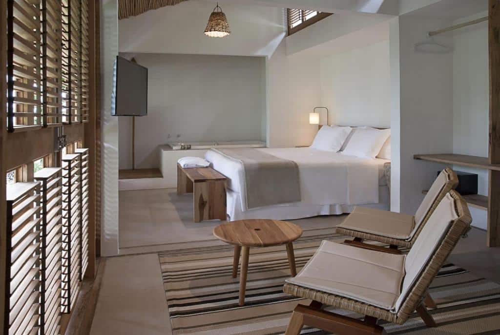 Quarto na Casa Mar Paraty com uma cama de casal, uma banheira, uma televisão, uma ampla sacada e duas cadeiras, tudo em tons de madeira e decoração rústica.