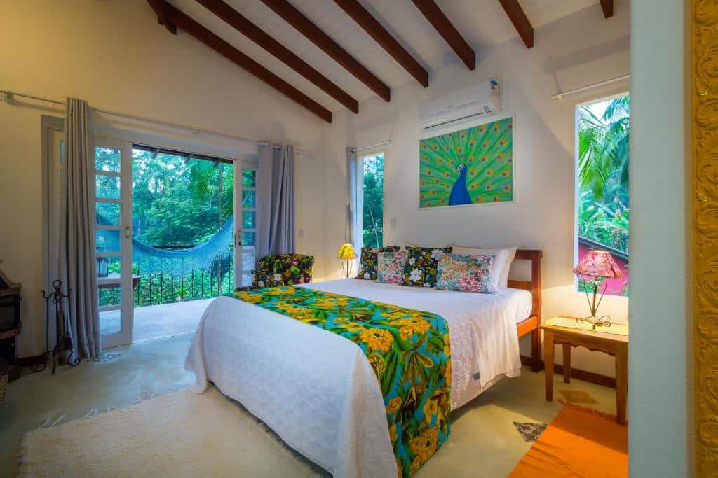 Quarto amplo no Pouso Vivenda da Mata com uma varanda com rede, uma cama de casal, decoração de flores, um ar-condicionado e um tapete branco, para representar hotéis em Paraty.