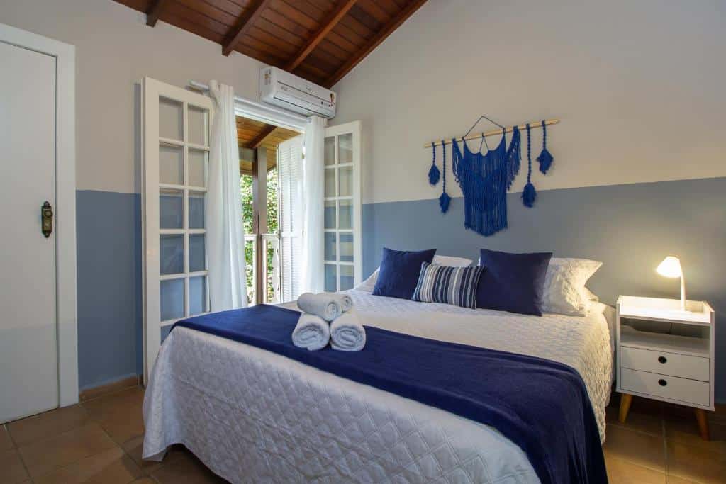 Quarto da Estrela do Mar Paraty com tudo decorado em branco e azul, a cama de casal é coberta por toalhas, travesseiros e almofadas, há uma sacada e um banheiro, assim como um pequeno móvel com um abajur do lado da cama.