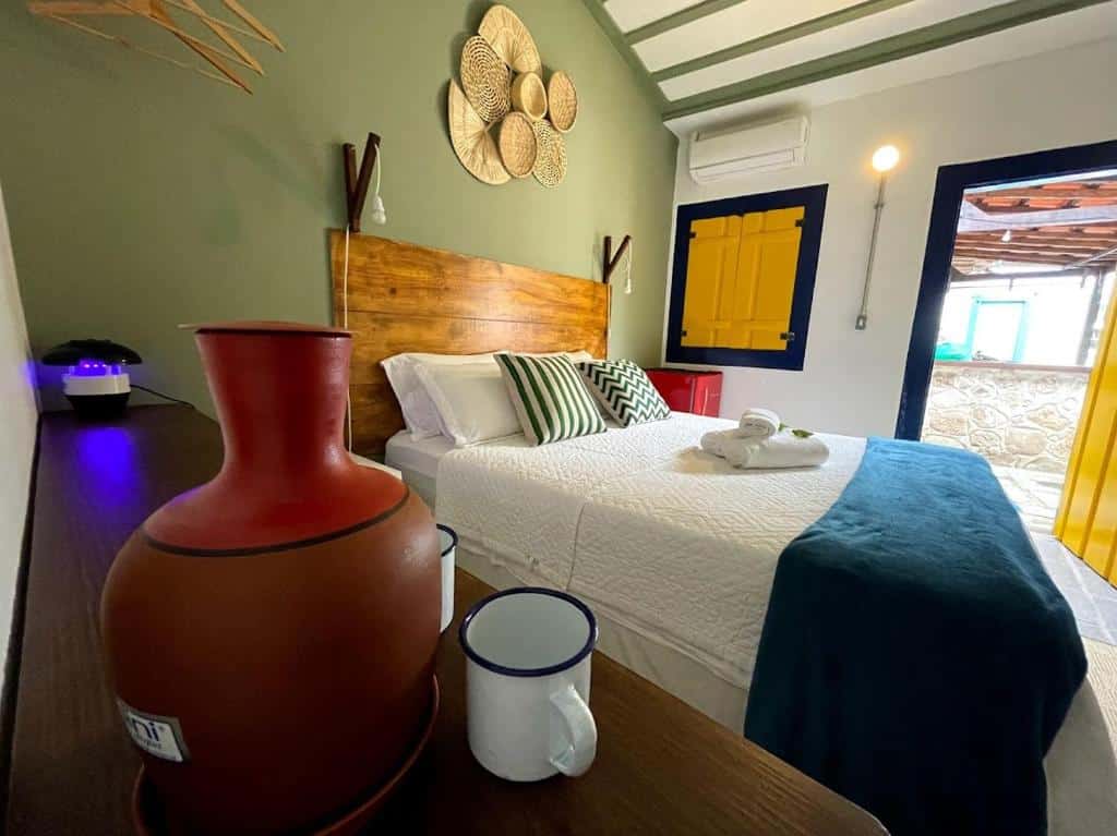 Quarto nas Casas do Pátio Pousada & Bar com uma decoração colonial, uma cama de casal, um frigobar, um ar-condicionado, uma cômoda com uma moringa com duas canecas, tudo em tons de azul, branco e azul, para representar hotéis em Paraty.