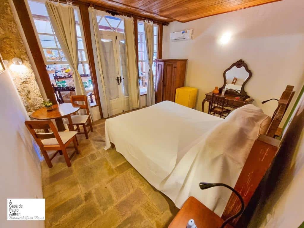 Quarto na A Casa de Paulo Autran com uma cama de casal, um frigobar, um armário, um ar-condicionado, uma penteadeira com uma cadeira, uma pequena mesa com duas cadeiras, tudo decorado em estilo colonial, com uma coluna de pedra e uma ampla porta que leva até a sacada.
