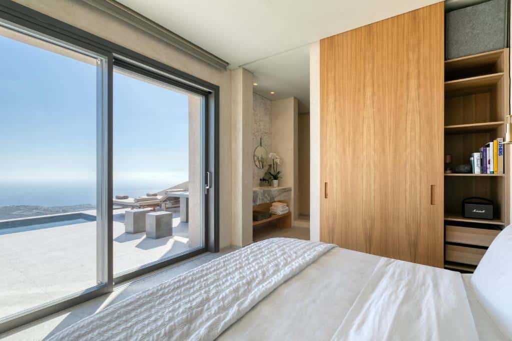 Quarto moderno com armário de madeira, roupa de cama branca e uma grande janela com vista para a varanda e para o mar.