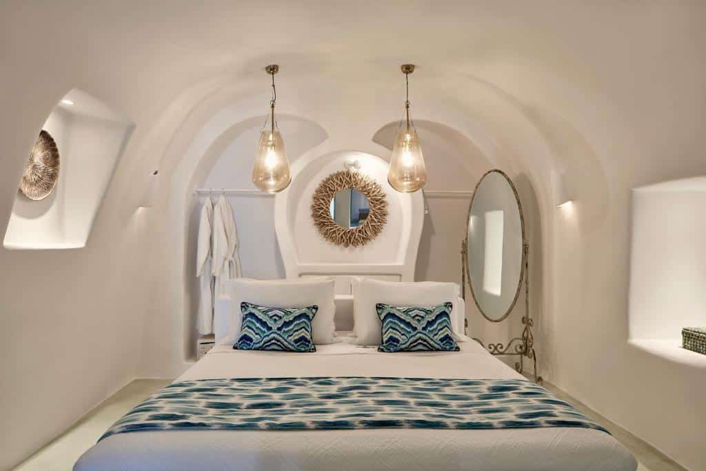 Um quarto branco em arco com uma cama de casal, travesseiros e tapete com estampa azul, duas luzes pendentes e um grande espelho oval.