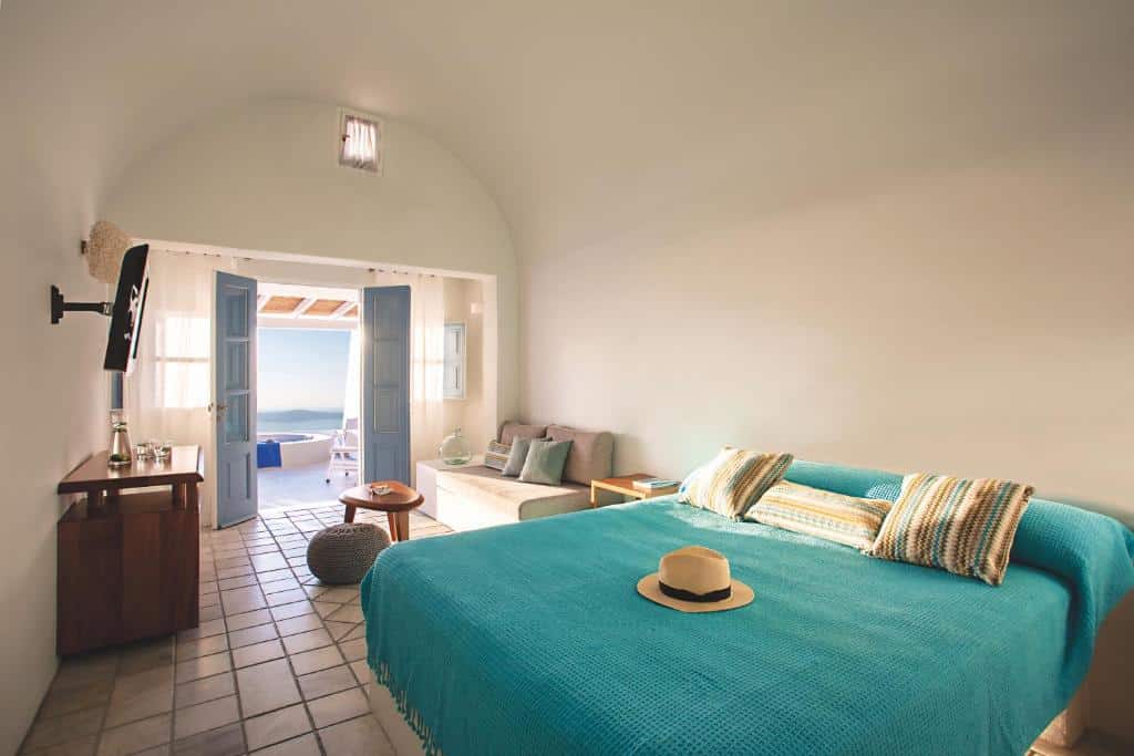 Um quarto bem iluminado com uma cama de casal, roupa de cama azul, um sofá, portas duplas abertas e vista para o mar do lado de fora.
