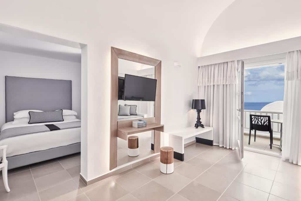 Quarto de hotel minimalista com cama, TV, escrivaninha e varanda com vista para o oceano sob um céu nublado.