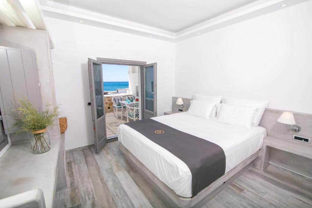 Quarto de hotel bem iluminado com uma cama de casal, detalhes em cinza e portas abertas que levam a uma varanda com vista para o mar.