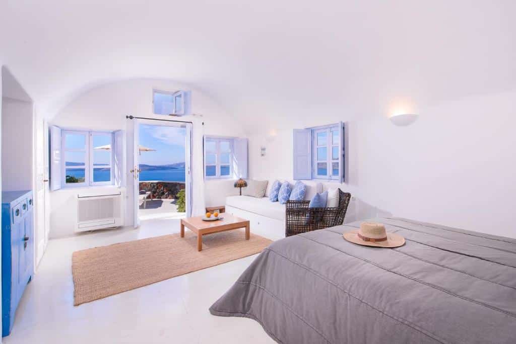 Quarto branco brilhante com detalhes em azul, janelas abertas e porta para um pátio com vista para o mar, sofá e mesa de centro.
