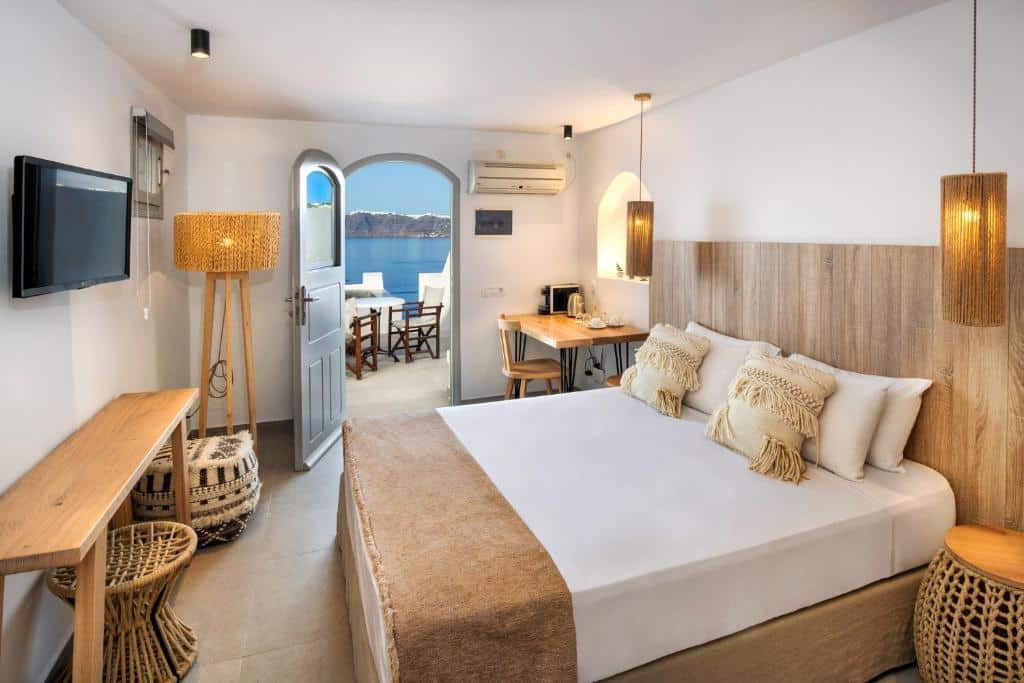 Um quarto de hotel moderno com uma cama de casal, escrivaninha, TV e porta aberta que dá para uma varanda com vista para o mar.