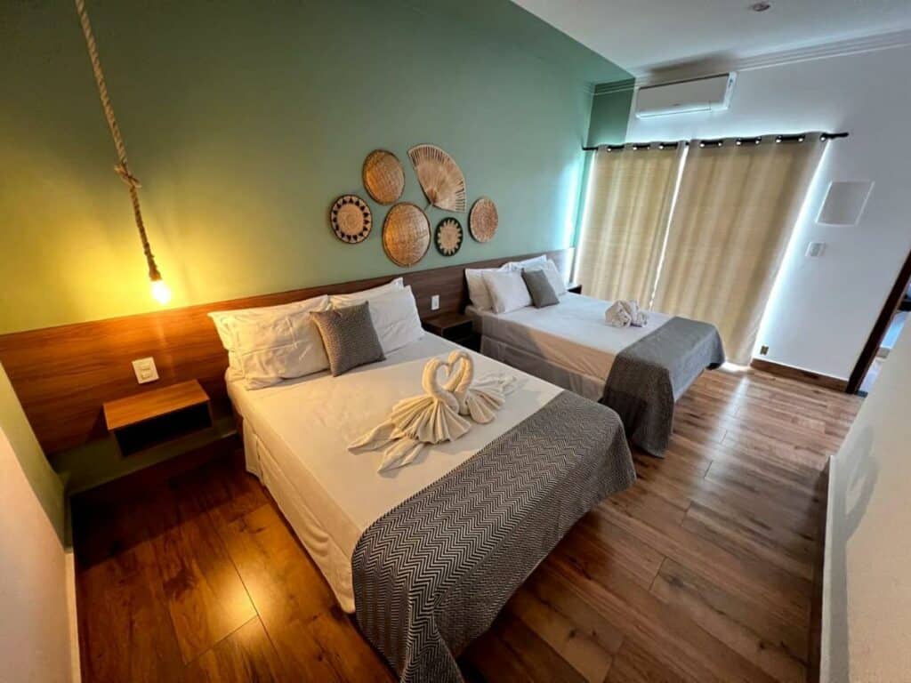 Quarto no Hotel Fazenda Vale da Cachoeira com uma cama de casal, uma cama de solteiro, uma cabeceira, um ar-condicionado, uma varanda com cortinas, o chão é de madeira e a decoração é rústica, para representar hotéis em Serra Negra