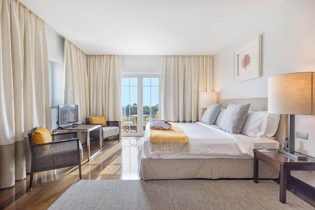 Quarto no The Lake Resort em Vilamoura, bem iluminado com uma cama de casal, poltrona, escrivaninha, TV, janela grande com cortinas e decoração neutra, para representar hotéis em Algarve.