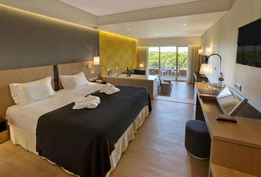 Quarto moderno no Falesia Hotel – Adults Only em Albufeira, com uma cama grande, escrivaninha, TV, área de estar e varanda com vista para a vegetação à luz do dia, para representar hotéis em Algarve.