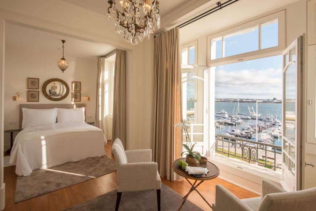 Quarto elegante no Grand House – Relais & Chateaux em Vila Real de Santo Antônio, com uma cama, cadeiras, lustre e janelas grandes com vista para a marina e a água, para representar hotéis em Algarve.