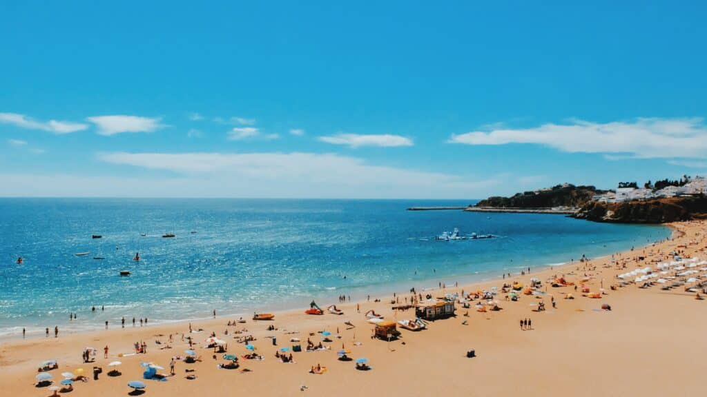 Praia de Albufeira em Algarve, de areia lotada com pessoas, guarda-sóis e mar azul sob um céu claro; falésias e edifícios distantes visíveis.