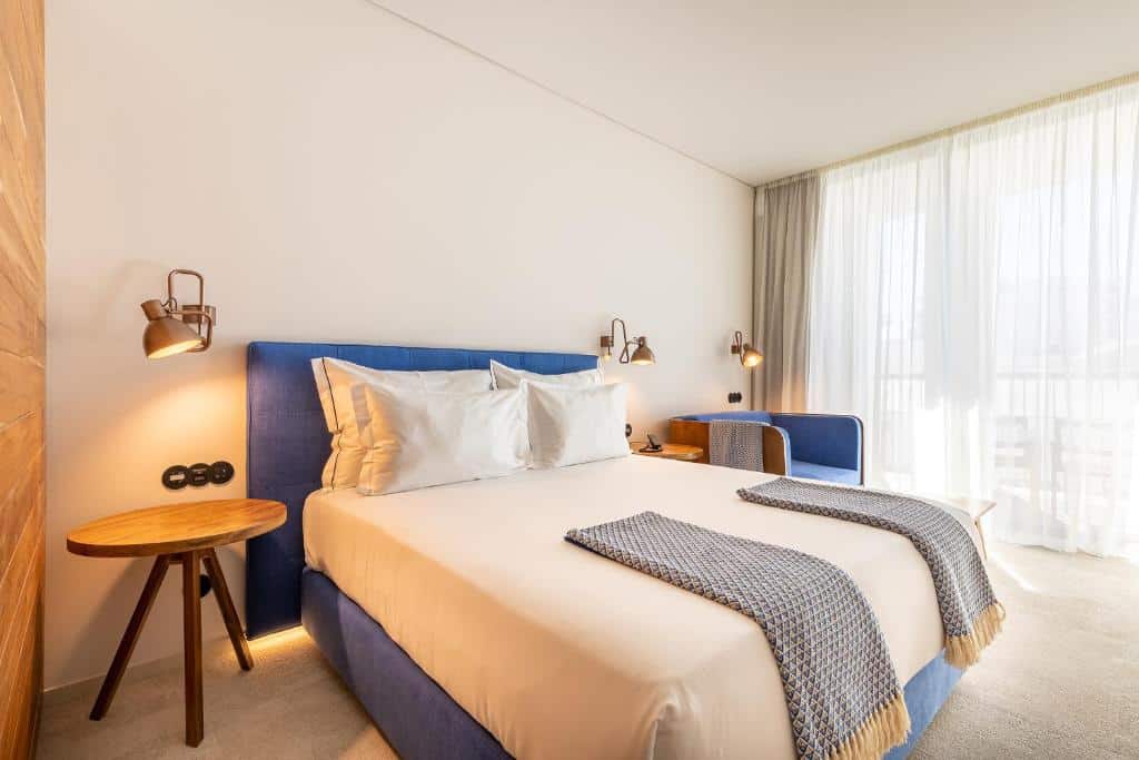 Quarto moderno no 3 HB Faro, em Faro, com uma cama estofada azul, roupa de cama branca, duas mantas cinza e grandes cortinas transparentes que deixam entrar a luz, para representar hotéis em Algarve.