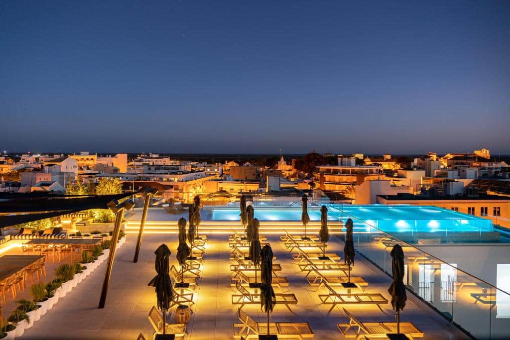 Piscina na cobertura e área de lounge com espreguiçadeiras vazias à noite, com vista para a paisagem urbana sob um céu claro, na propriedade do 3HB Faro em Faro.