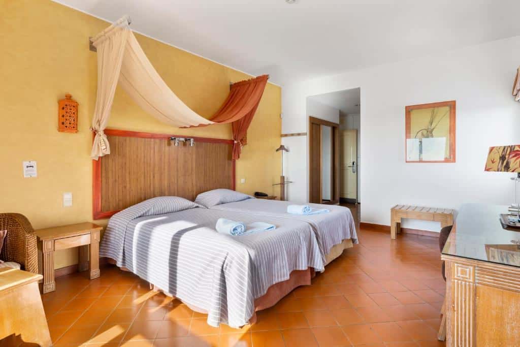 Quarto no Cerro da Marina Hotel em Albufeira, com duas camas de solteiro, roupa de cama listrada, um dossel e piso de azulejo; toalhas colocadas em cada cama, para representar hotéis em Algarve.