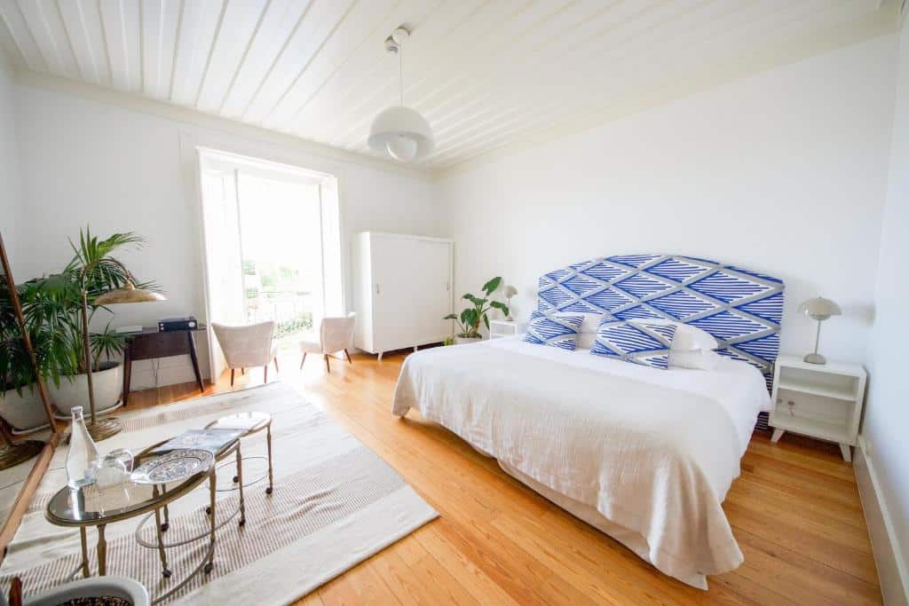 Quarto bem iluminado com piso de madeira, cama de casal, cabeceira estampada, mesinhas, cadeiras e janela grande com luz natural, na hospedagem Casa Mãe em Lagos, para representar hotéis em Algarve.