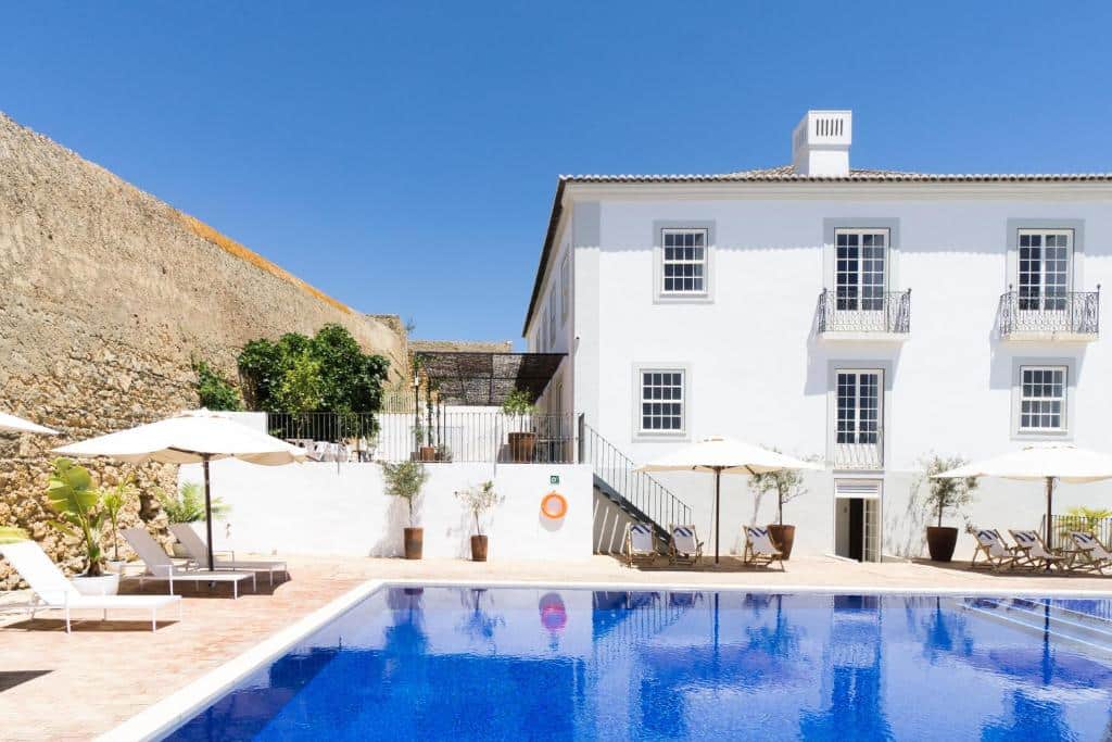 Uma piscina externa retangular com água azul fica em frente a um prédio branco de dois andares com varandas e guarda-sóis à beira da piscina, na propriedade da Casa Mâe em Lagos.