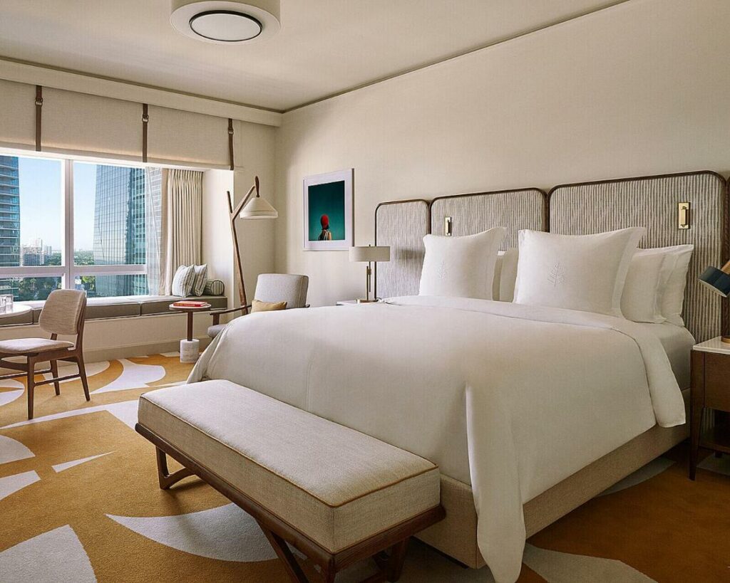 Quarto do Four Seasons Hotel Miami com cama de casal do lado direito da imagem, no pé da cama um banco estofado e do lado direito uma cômoda com luminária, ainda do lado esquerdo uma poltrona. Representa hotéis para Brasileiros em Miami.
