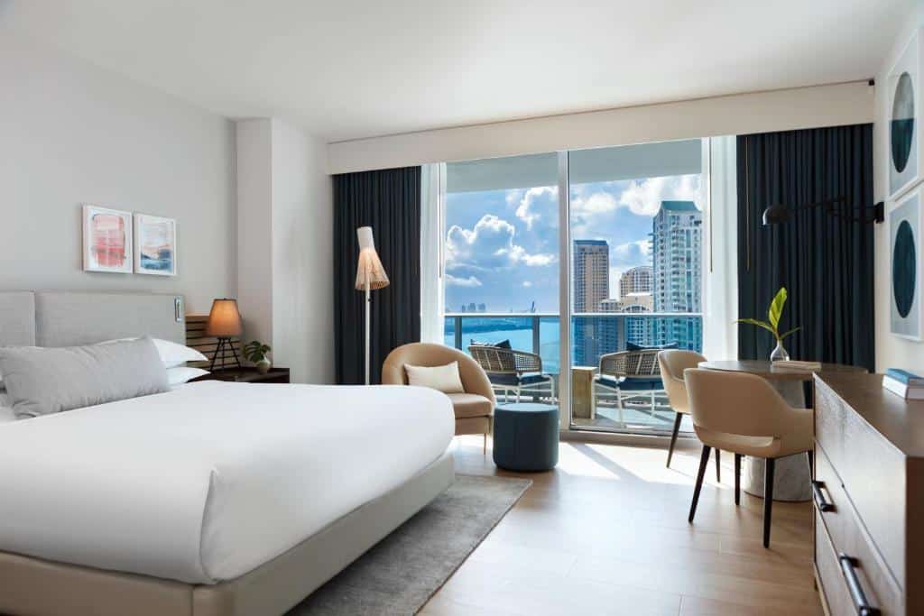 Quarto do Kimpton EPIC Hotel, an IHG Hotel com cama de casal do lado esquerdo da imagem, uma cômoda do lado esquerdo da cama em frente a cama uma cômoda do lado esquerdo uma mesa com duas cadeiras. Representa hotéis para Brasileiros em Miami.