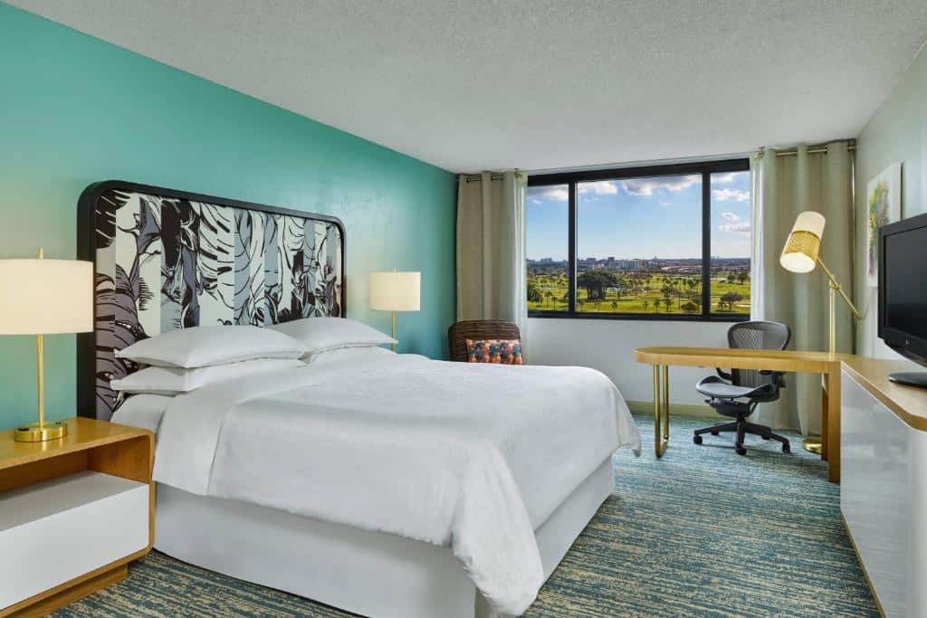 Quarto do Sheraton Miami Airport Hotel and Executive Meeting Center com cama de casal do lado esquerdo da imagem com uma cômoda em cada lado da cama com luminária, em frente a cama uma cômoda com TV e do lado esquerdo da cômoda uma mesa com cadeira. Representa hotéis para Brasileiros em Miami.